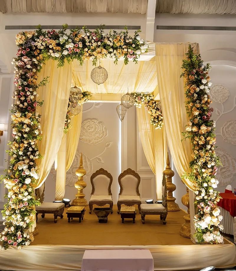 A floral mandap draped in cream fabric with two gilded chairs, low carved wooden stools and gold finials on a raised platform, in warm light