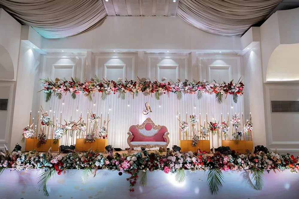 A floral stage in the hall with a pink and gold sofa, tall flower stands and a border of flowers and palm leaves along the front